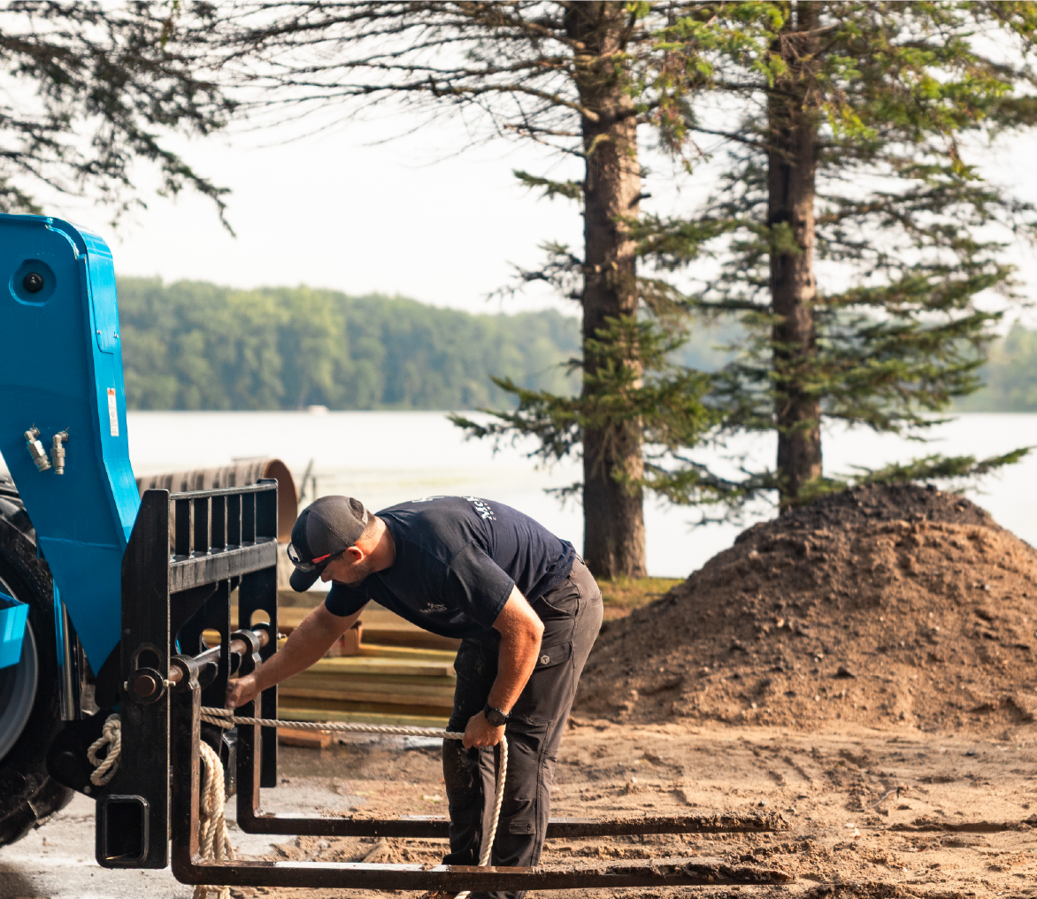Man wearing a black cap and dark clothing adjusting a rope on a blue piece of heavy equipment outdoors near trees and a lake.