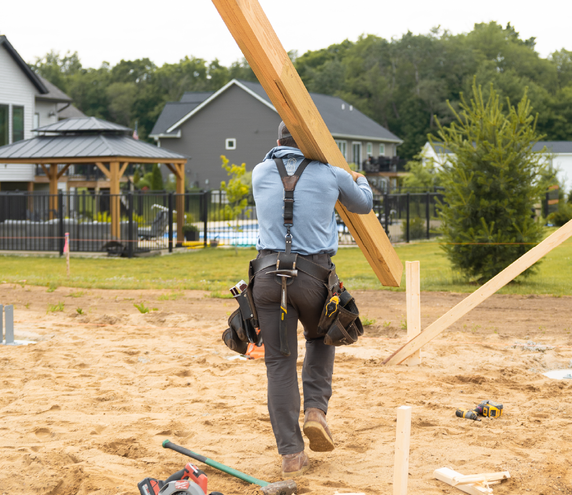 Construction worker carrying a wooden beam on a sandy site near houses and a gazebo.