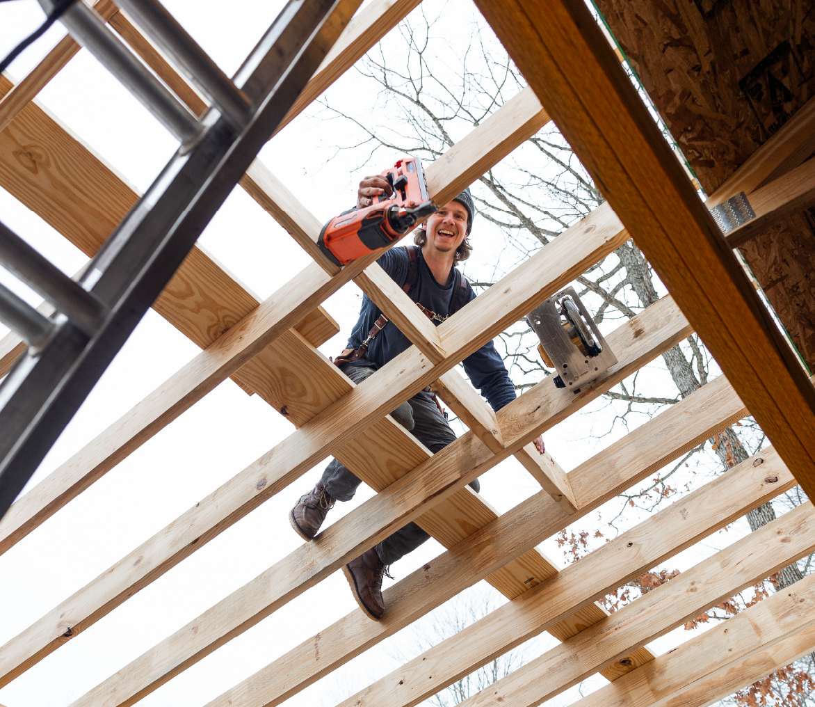 Construction worker standing on wooden beams holding a nail gun and smiling during roof framing.