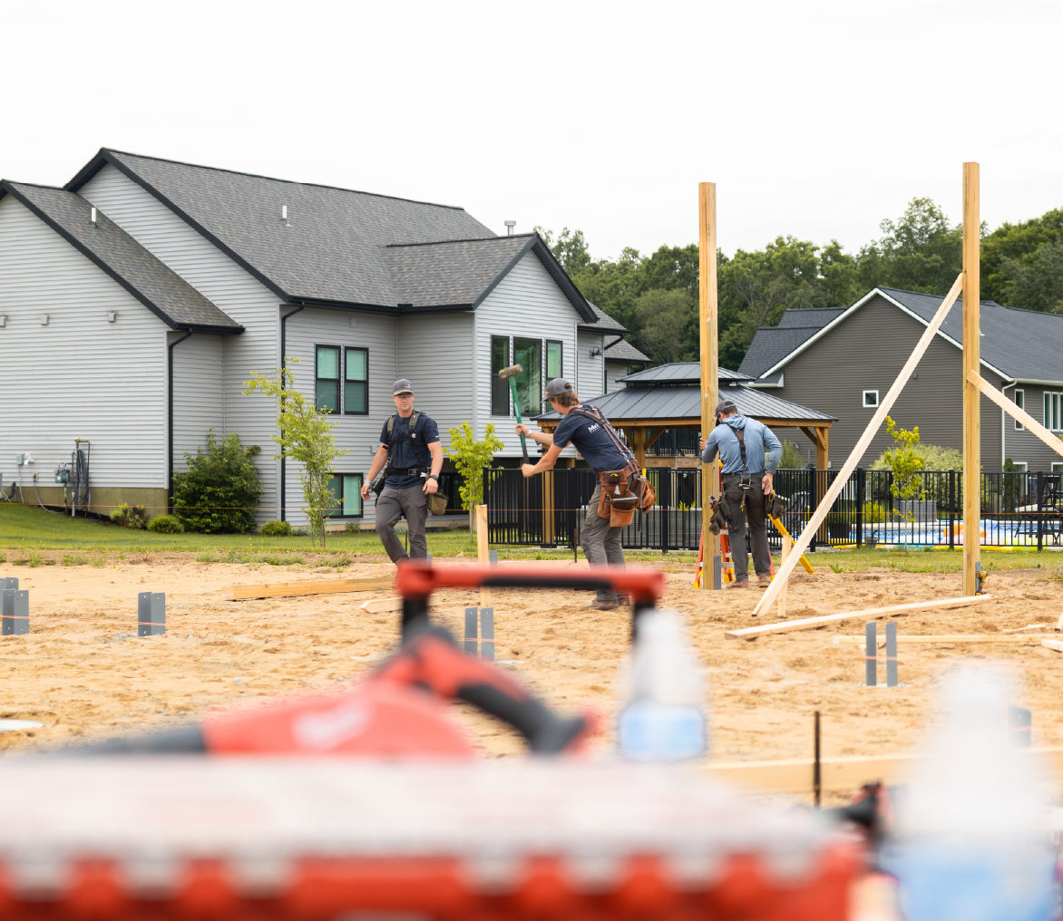 Three construction workers assembling wooden posts on a sandy site near residential houses.
