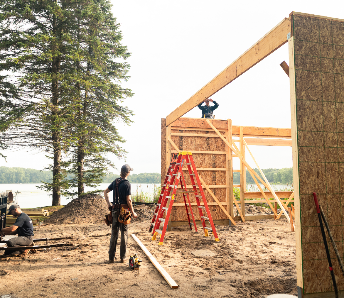 Three construction workers assembling wooden framing for a building near a lakeside with trees in the background.