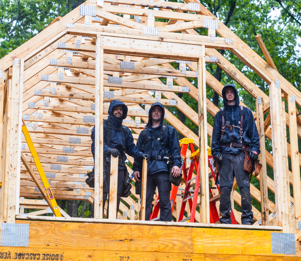 Three construction workers wearing black rain jackets and tool belts standing inside a wooden frame structure under construction with ladders visible behind them.