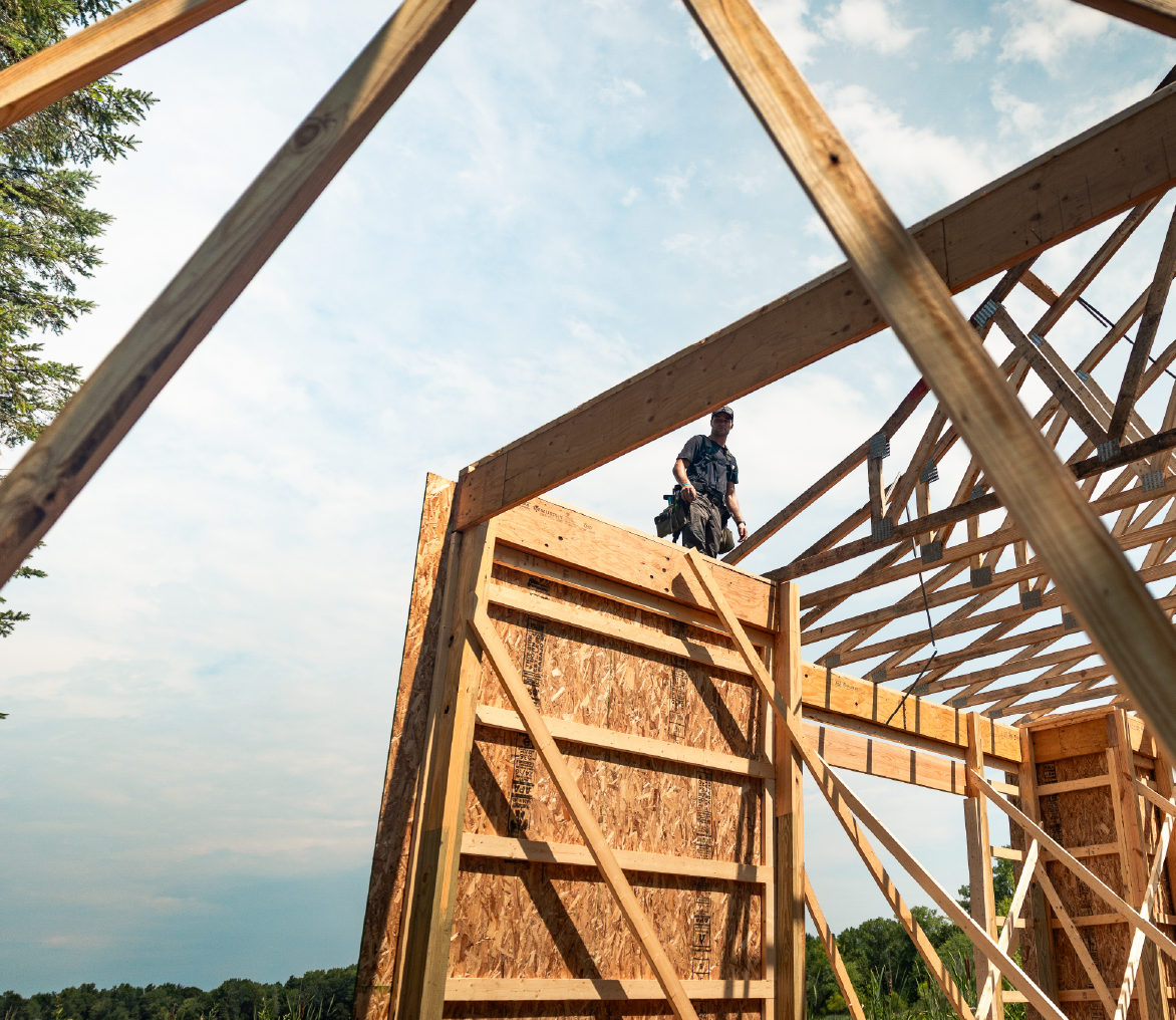 Construction worker standing on wooden framework of a house under a partly cloudy sky.