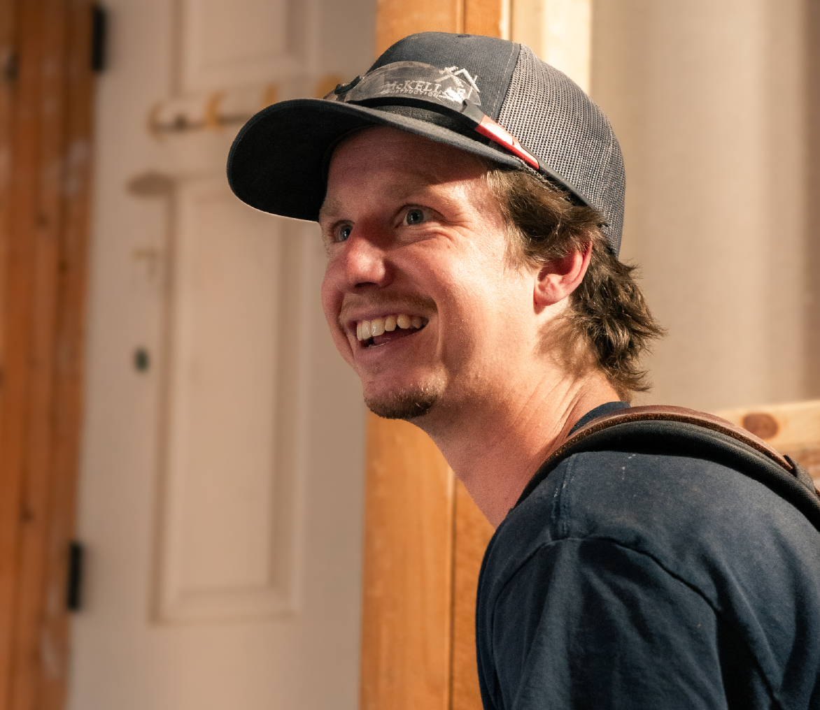 Smiling man wearing a black mesh baseball cap indoors with wooden walls and a white door in the background.