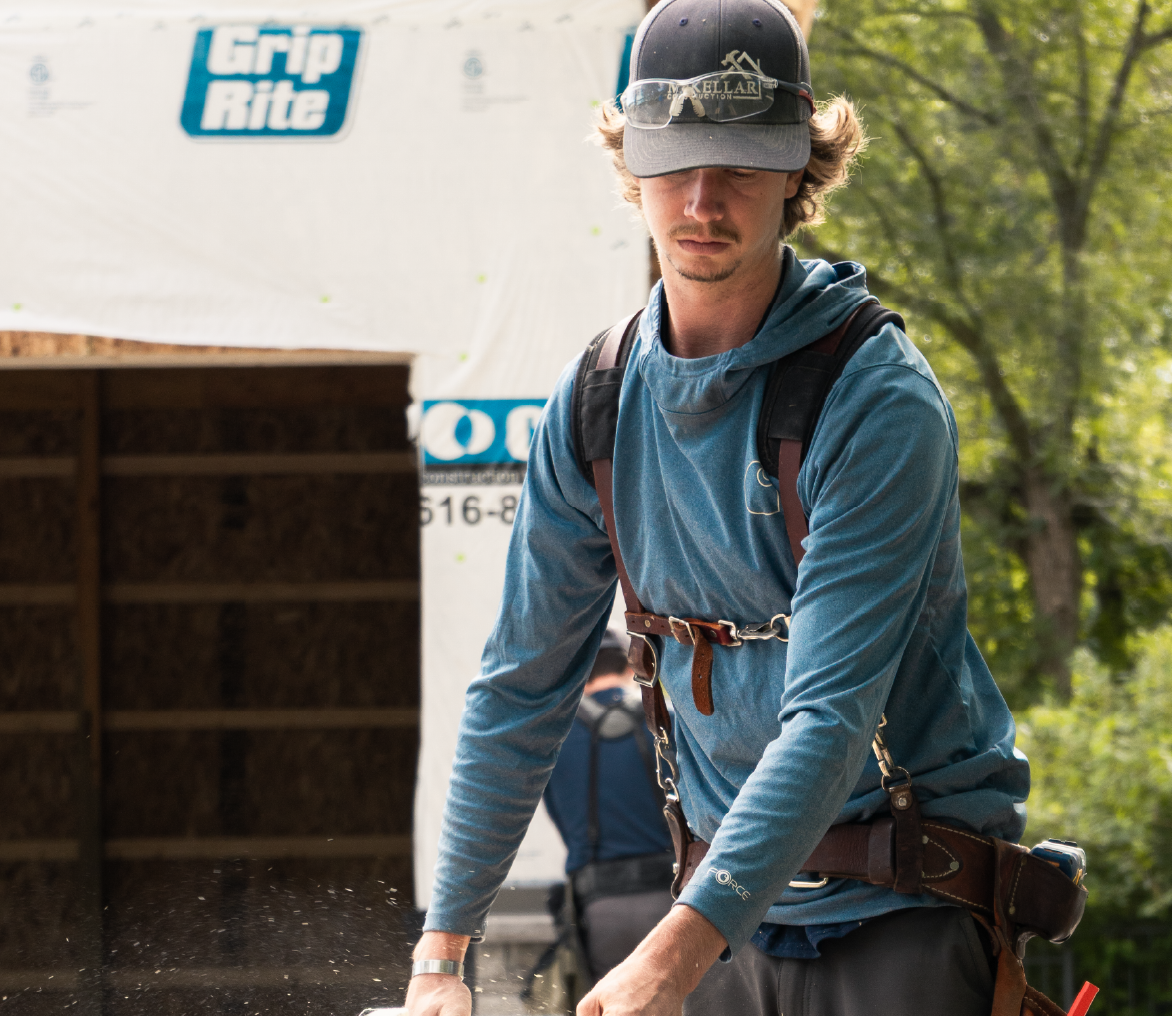 Construction worker wearing a cap and safety harness working outdoors near a building under construction.