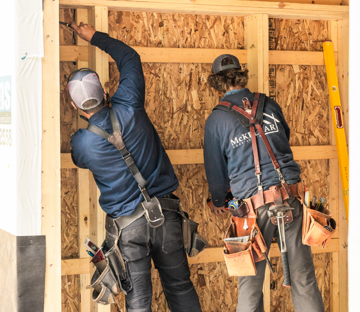 Two construction workers wearing tool belts and caps working on a wooden frame wall inside a building under construction.