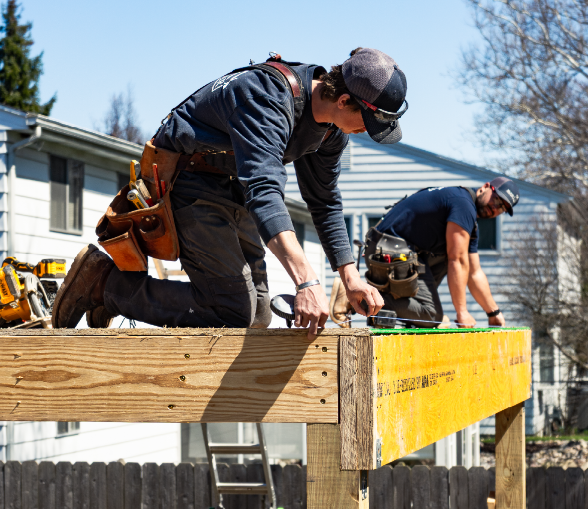 Two construction workers kneeling on a wooden platform, measuring and working on a framing project outdoors.