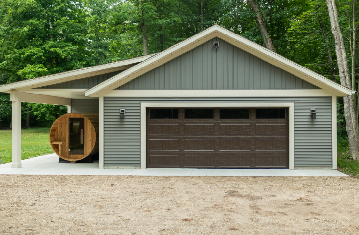 Gray detached garage with dark brown double garage door and a wooden barrel sauna on the side under a covered area, surrounded by trees.