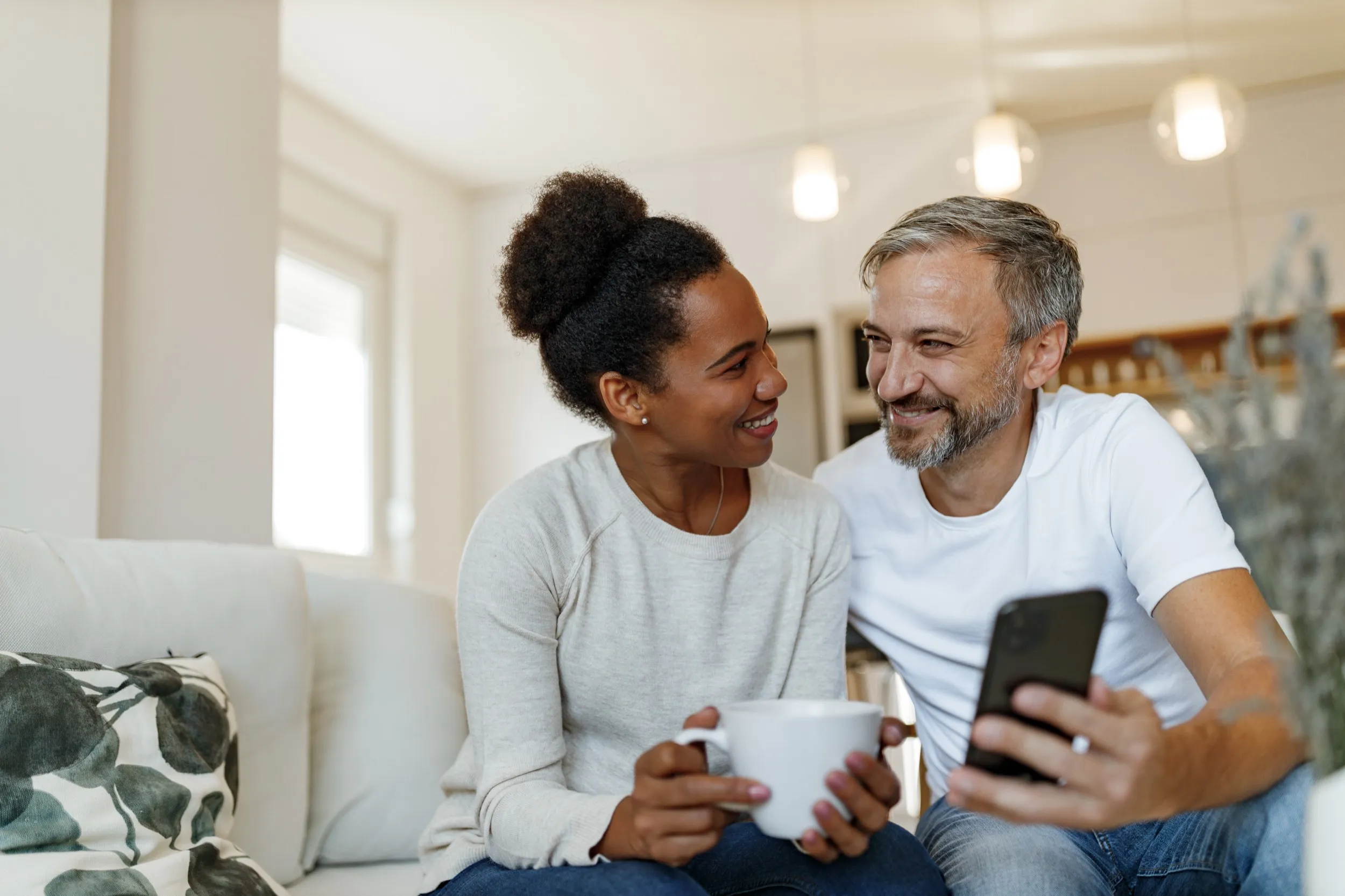 Two people smiling and sitting together on couch, holding coffee and phone