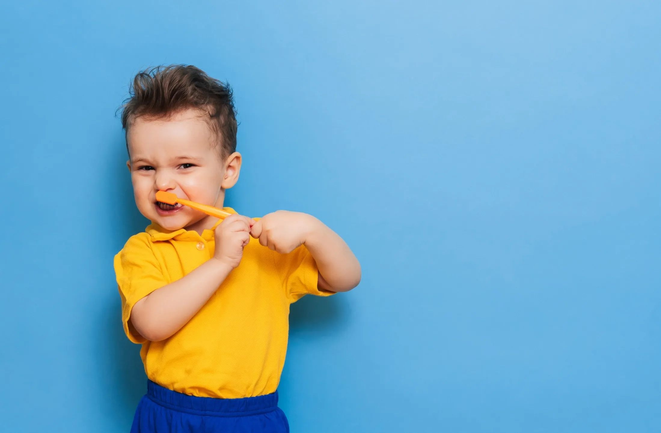 Young child in yellow shirt playfully biting yellow toothbrush against blue background