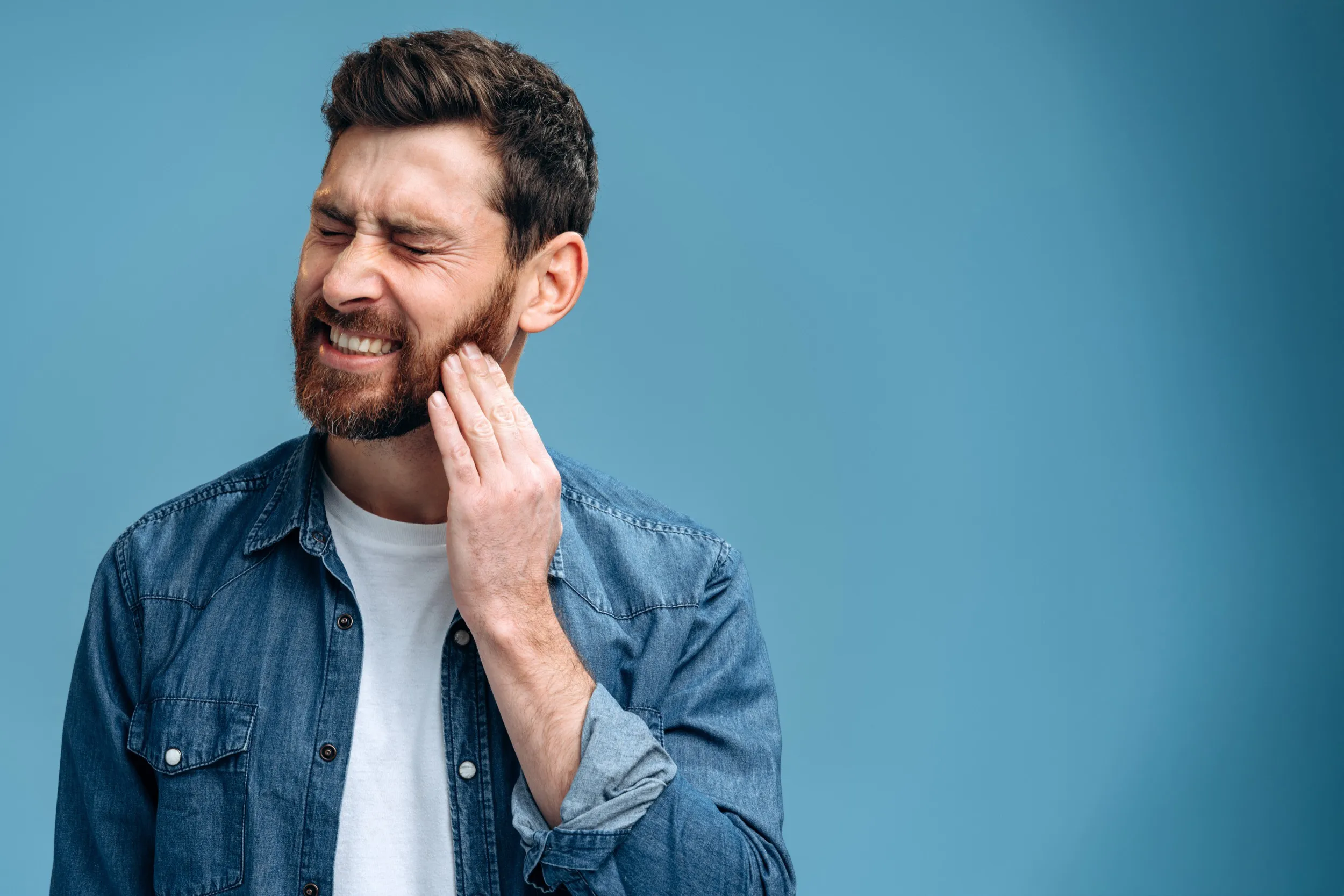 Bearded man in denim shirt grimacing and touching cheek in pain