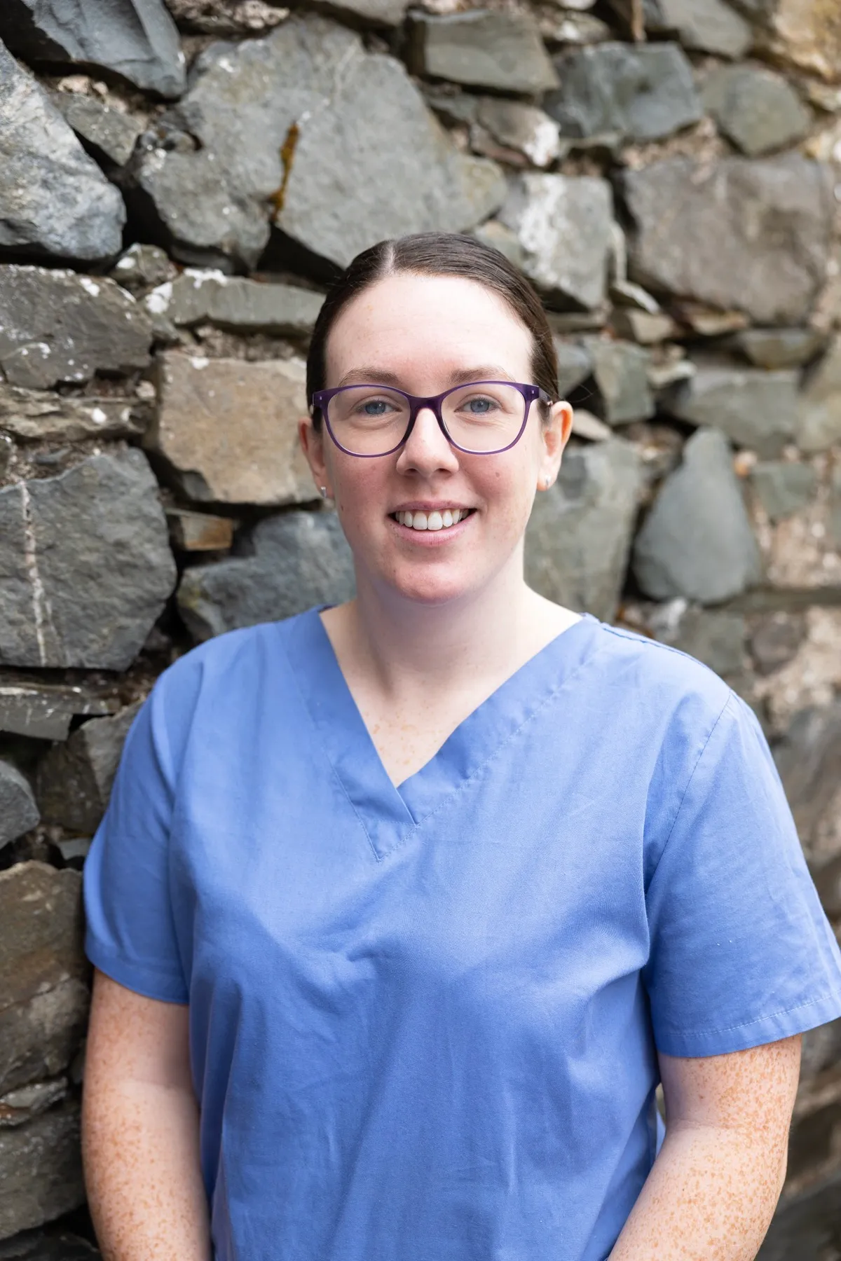 Smiling healthcare worker in blue scrubs standing against stone wall