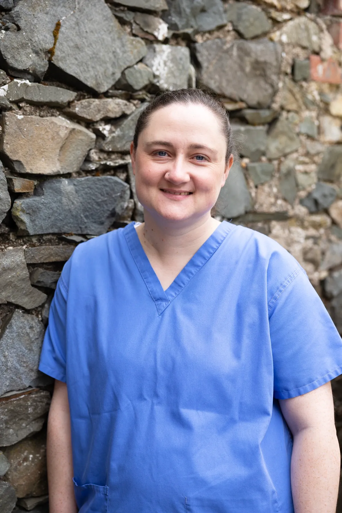 Smiling healthcare worker in blue scrubs standing against stone wall