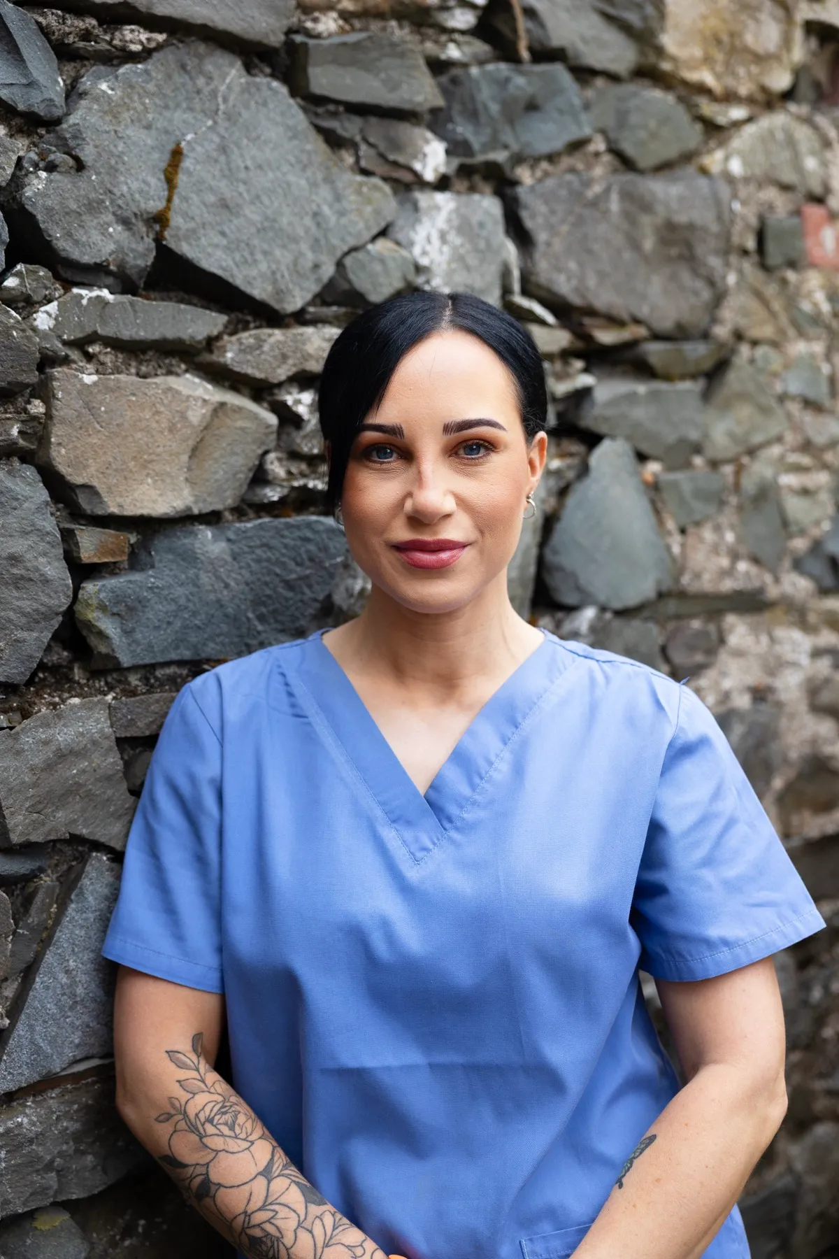Healthcare professional in blue scrubs standing against stone wall with tattoo