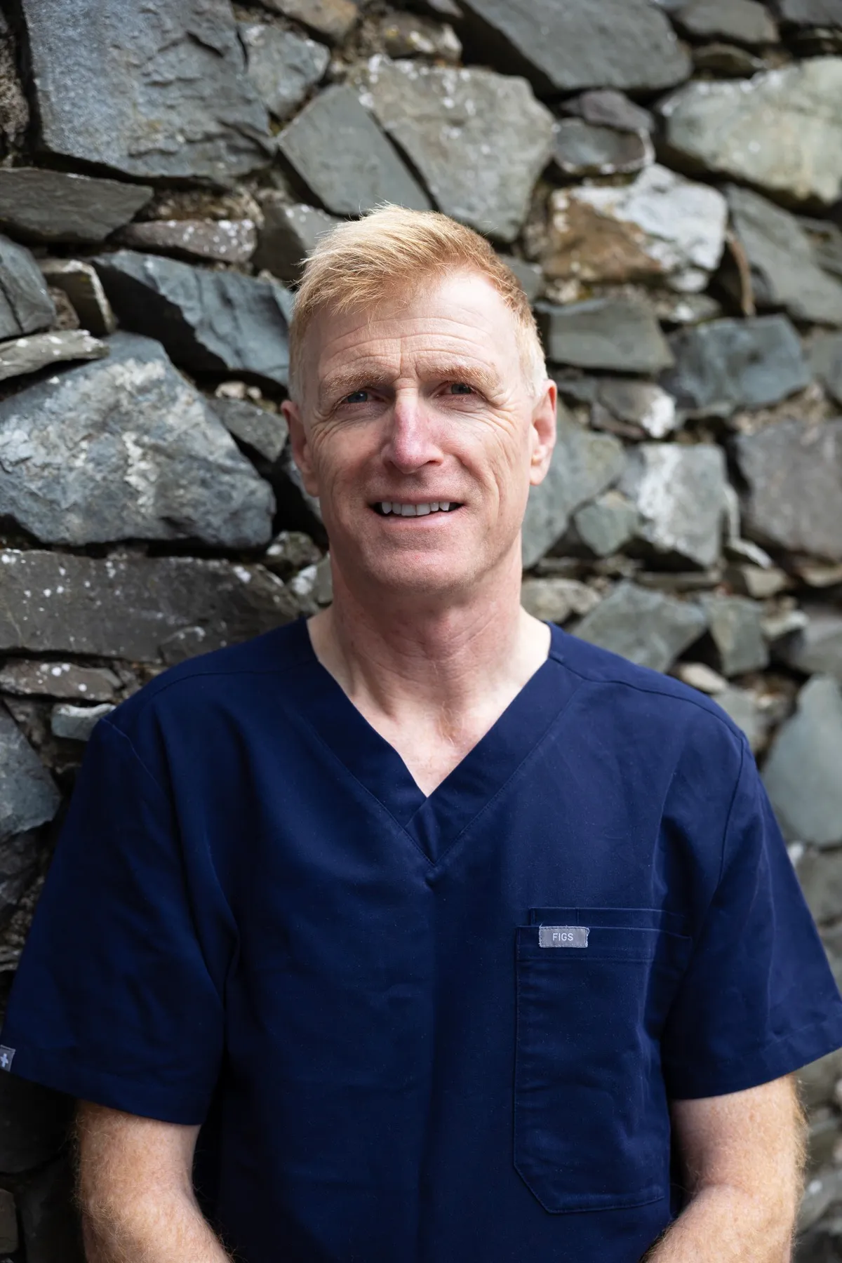 Smiling medical professional in blue scrubs standing against stone wall