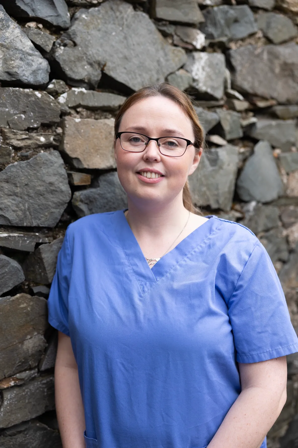 Smiling healthcare worker in blue scrubs standing against stone wall