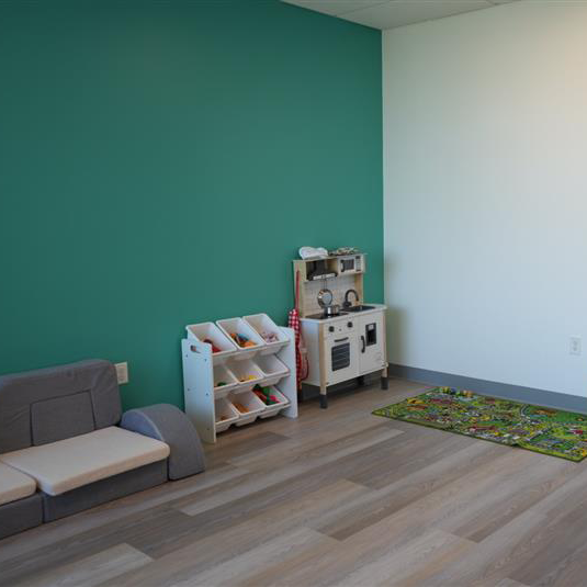 Play therapy room with a teal accent wall, modular soft seating, a toy shelf with bins, a play kitchen, and a car-themed play mat.