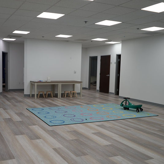 Open therapy room with a colorful circle-patterned learning rug, activity tables, wood stools, and a green ride-on toy.
