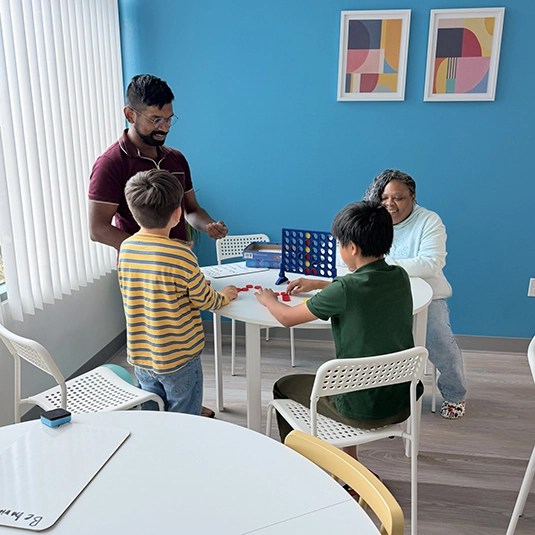 Therapy room with staff guiding two children through games and learning activities at a round table in front of a bright blue accent wall.