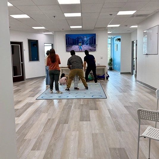 Open clinic activity area with families and staff gathered around a wall-mounted screen, ride-on toys, and a colorful learning rug.