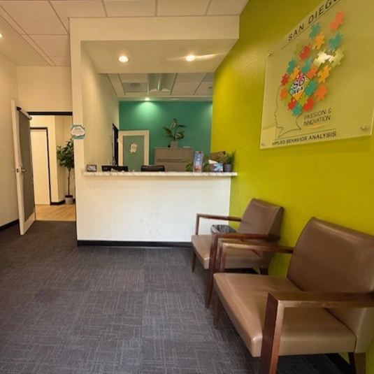 Welcoming reception and waiting area with comfortable chairs, a service counter, and a wall sign reading San Diego Passion and Innovation Applied Behavior Analysis.