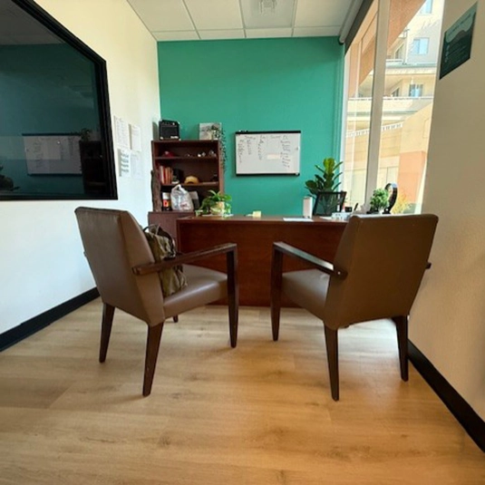 Private consultation office with a desk, two leather guest chairs, a teal accent wall, whiteboard, and an observation window.