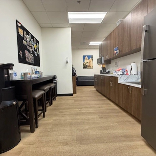 Staff kitchen and break area with brown wood cabinetry, a refrigerator, bar stools at a counter, and a bulletin board on the wall.