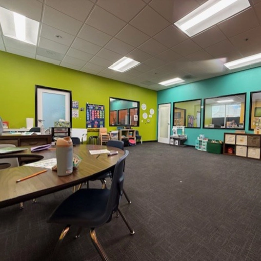 Spacious therapy room with bright green and teal accent walls, learning boards, tables and chairs, and educational materials for group sessions.