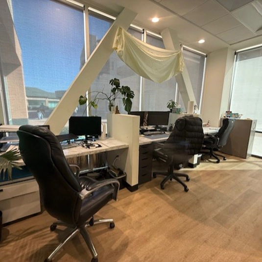 Office workspace with multiple desks, computer monitors, ergonomic chairs, and natural light from large windows.