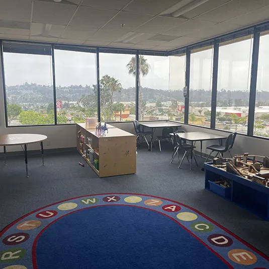 Sunlit therapy room at the Oceanside ABA clinic with child-sized tables, an alphabet circle rug, learning shelves, and windows overlooking the city.