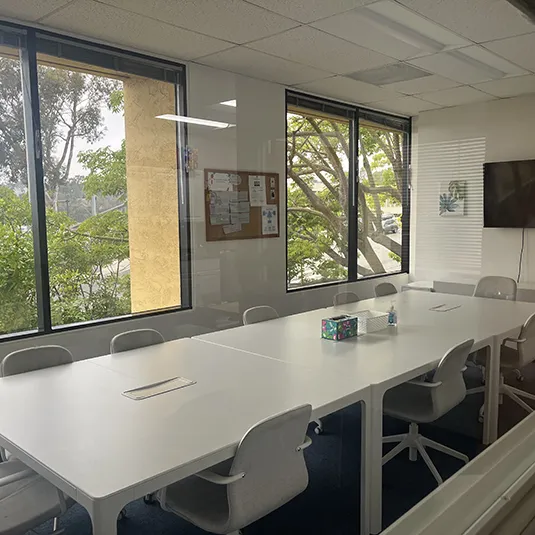 Conference room with a long white meeting table, gray chairs, a wall-mounted display screen, and large windows framing leafy trees.