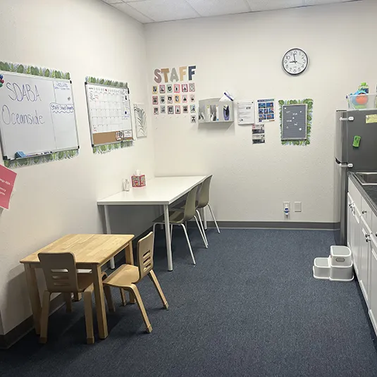 Therapy and staff room with a child-sized wooden table, scheduling and staff boards, a wall clock, and a kitchenette with a step stool.