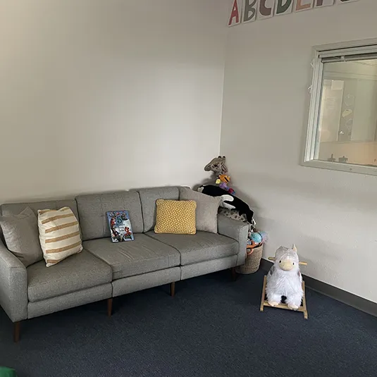 Comfortable family waiting area with a gray sofa, plush toys, children's books, an alphabet wall border, and an interior observation window.