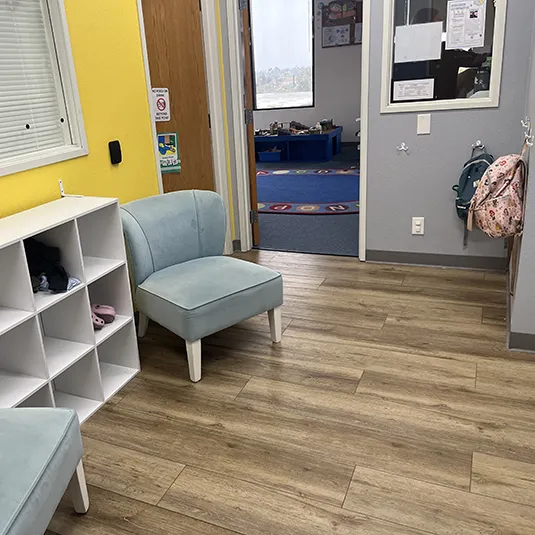Bright entry area with cubby storage, soft blue chairs, wall hooks for backpacks, and a doorway into a colorful therapy room with an alphabet rug.