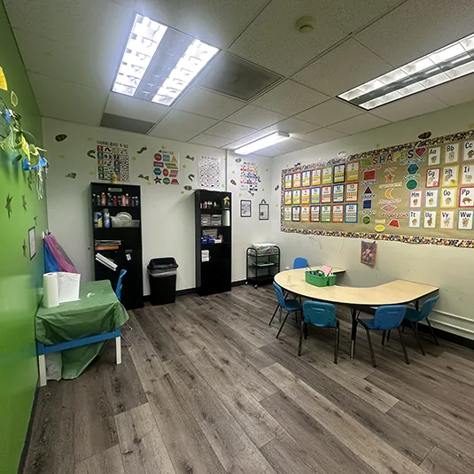 Bright therapy room with a green accent wall, a half-moon activity table with blue chairs, storage shelves, and alphabet and shapes learning posters.