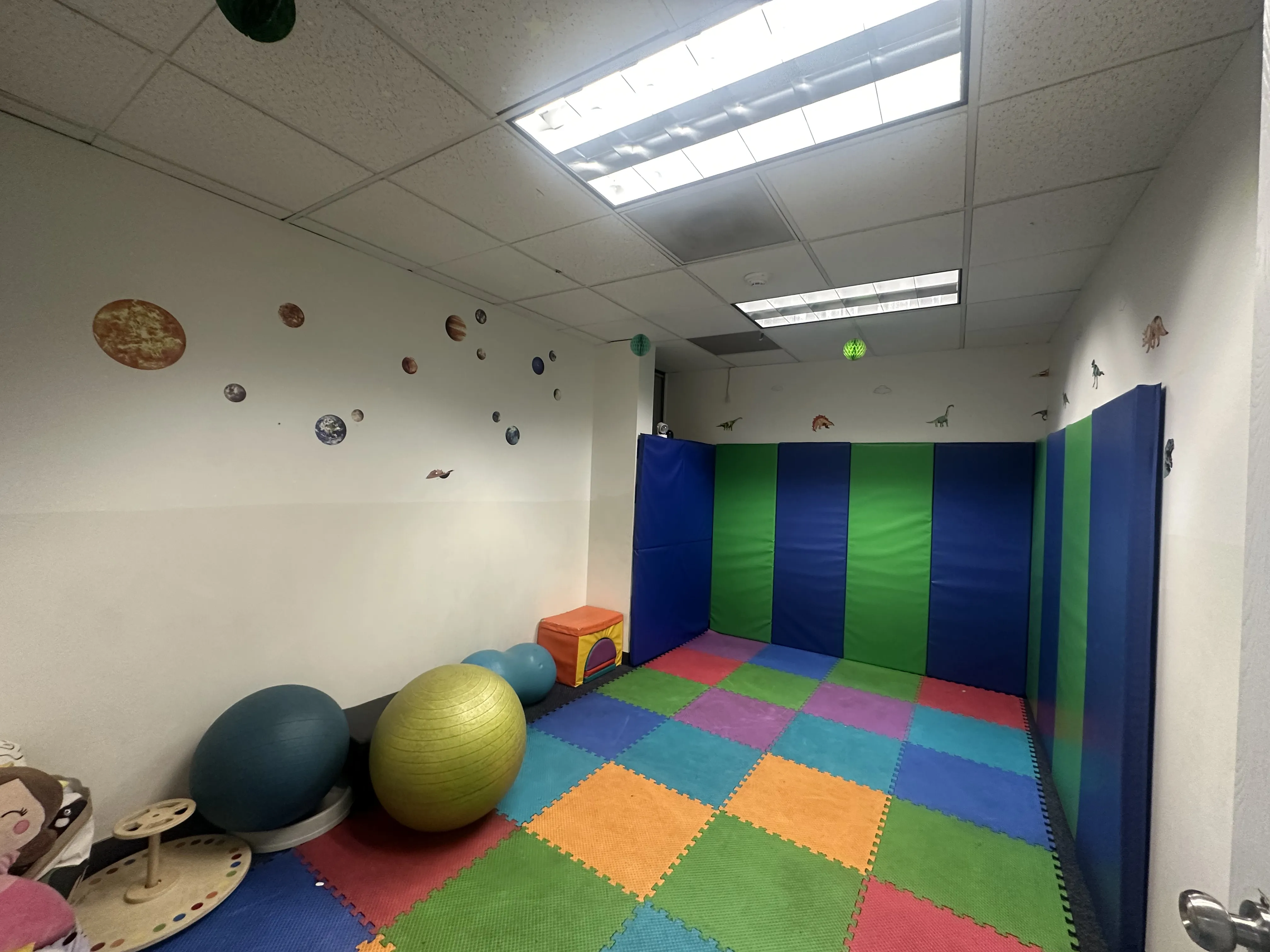 Padded sensory motor room with wall mats, therapy balls, a balance board, foam floor tiles, and a planets-and-dinosaurs wall theme.