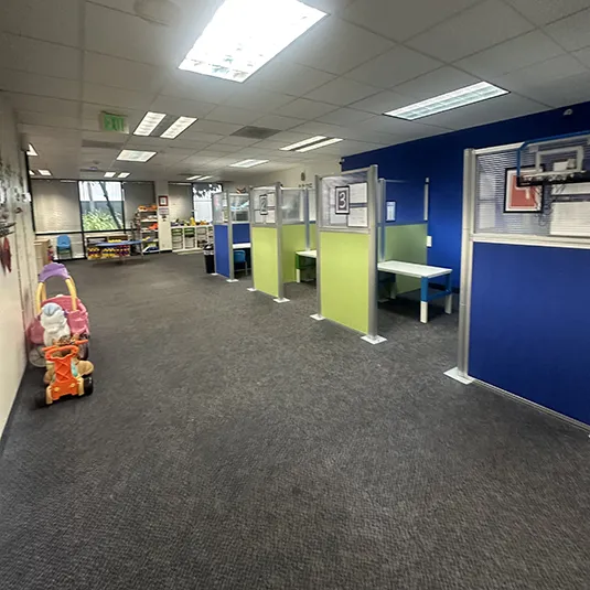 Open therapy area with individual partitioned workstations, child-sized desks and chairs, a basketball hoop, and a play space along the windows.