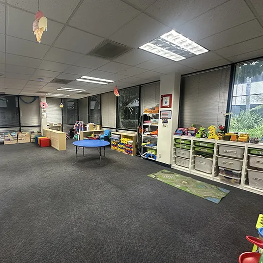 Spacious therapy room with low toy shelves, learning materials, a round blue activity table, and large windows with blinds for natural light.