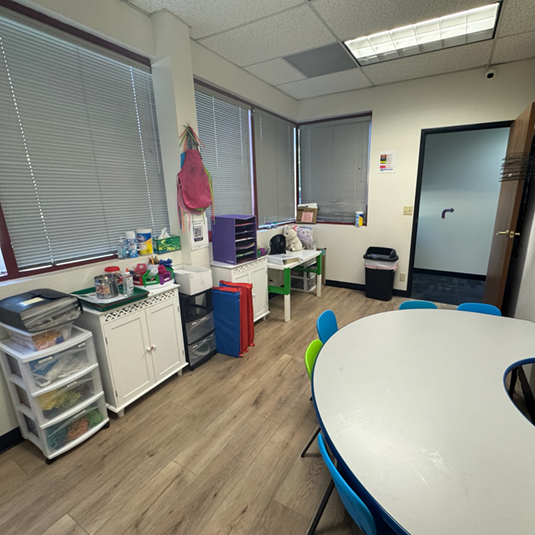 Therapy session room with a round activity table, blue and green chairs, organized supply storage, and windows with natural light.