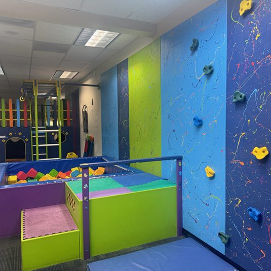 Sensory gym at the Lancaster ABA clinic with a climbing wall, ball pit, padded platforms, and a play structure in bright blues and greens.