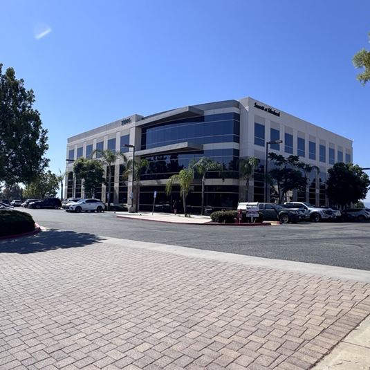 Exterior of the Temecula Valley clinic, a modern multi-story office building with palm trees and a paved parking area under a clear blue sky.
