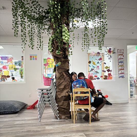 Open therapy and activity room with a large decorative tree centerpiece, student artwork on the walls, and a child-sized table and chairs.