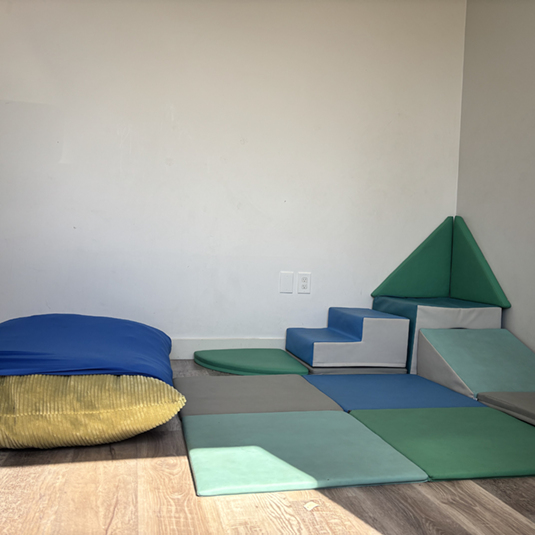 Soft play corner with padded foam climbing shapes, a step wedge, and floor cushions in blue and green for movement and sensory breaks.