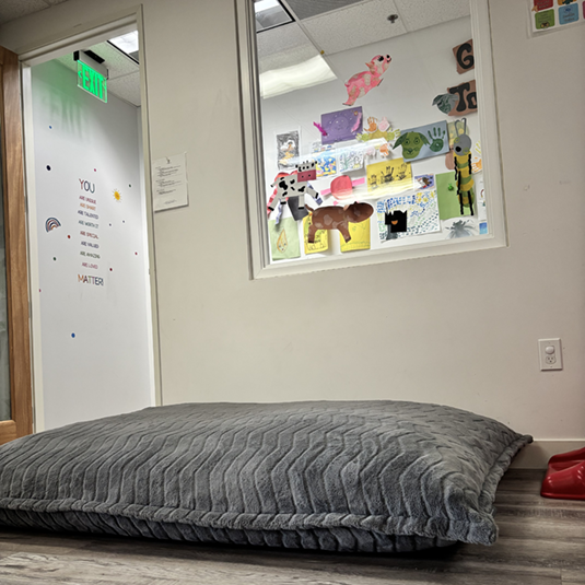 Quiet break area with a large floor cushion, an observation window into a decorated therapy room, and an affirmation decal on the wall.
