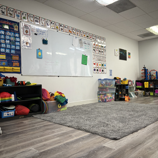 Structured learning room with a whiteboard, alphabet and calendar wall charts, visual supports, low storage shelves, and a soft area rug.