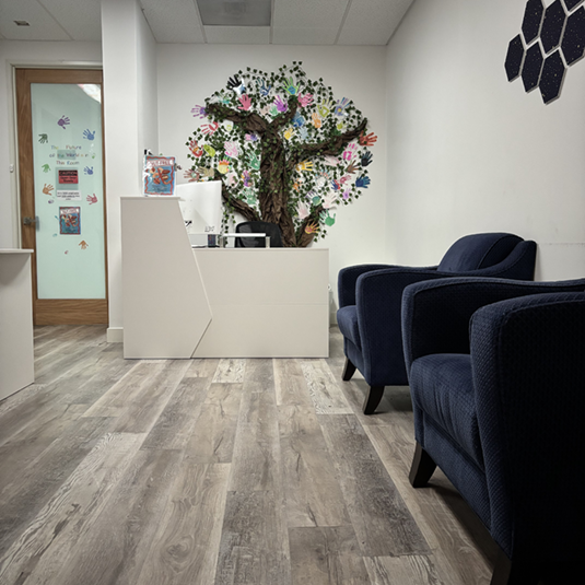 Reception area with a white welcome desk, a colorful handprint tree mural, navy waiting chairs, and light wood-look flooring.