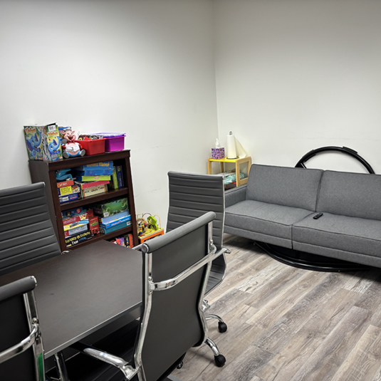 Consultation and meeting room with a conference table, office chairs, a bookshelf of games and learning materials, and a gray sofa.