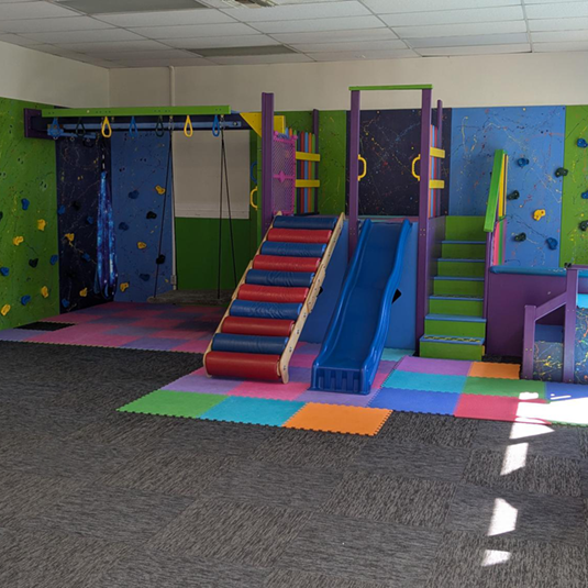 Indoor motor gym at the San Fernando clinic with climbing walls, hanging rings, a wooden staircase, a blue slide, and interlocking foam floor mats.