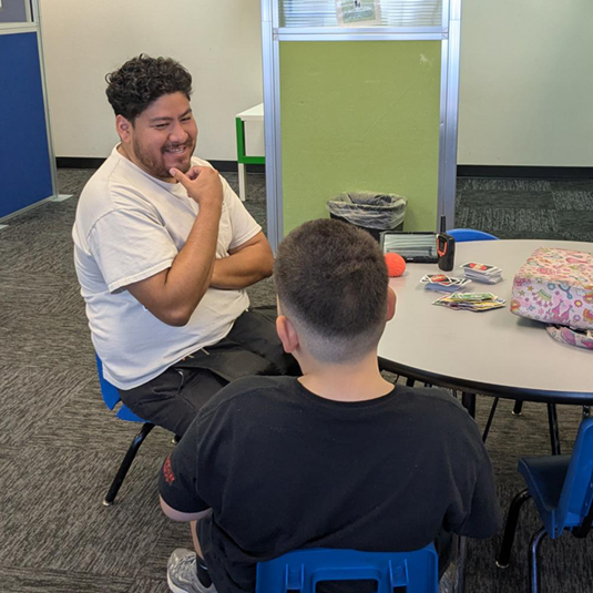 A one-on-one session at a round activity table in a carpeted learning room, with learning cards on the table and a green privacy partition behind.