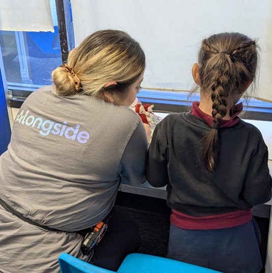 A clinician in an Alongside team shirt working one-on-one with a child at a learning desk beside a window.